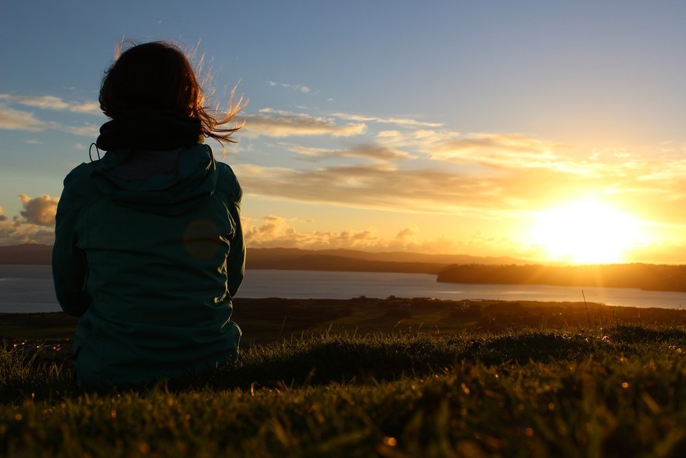 A person sitting in tall grass at golden hour, looking out over water, settled and present